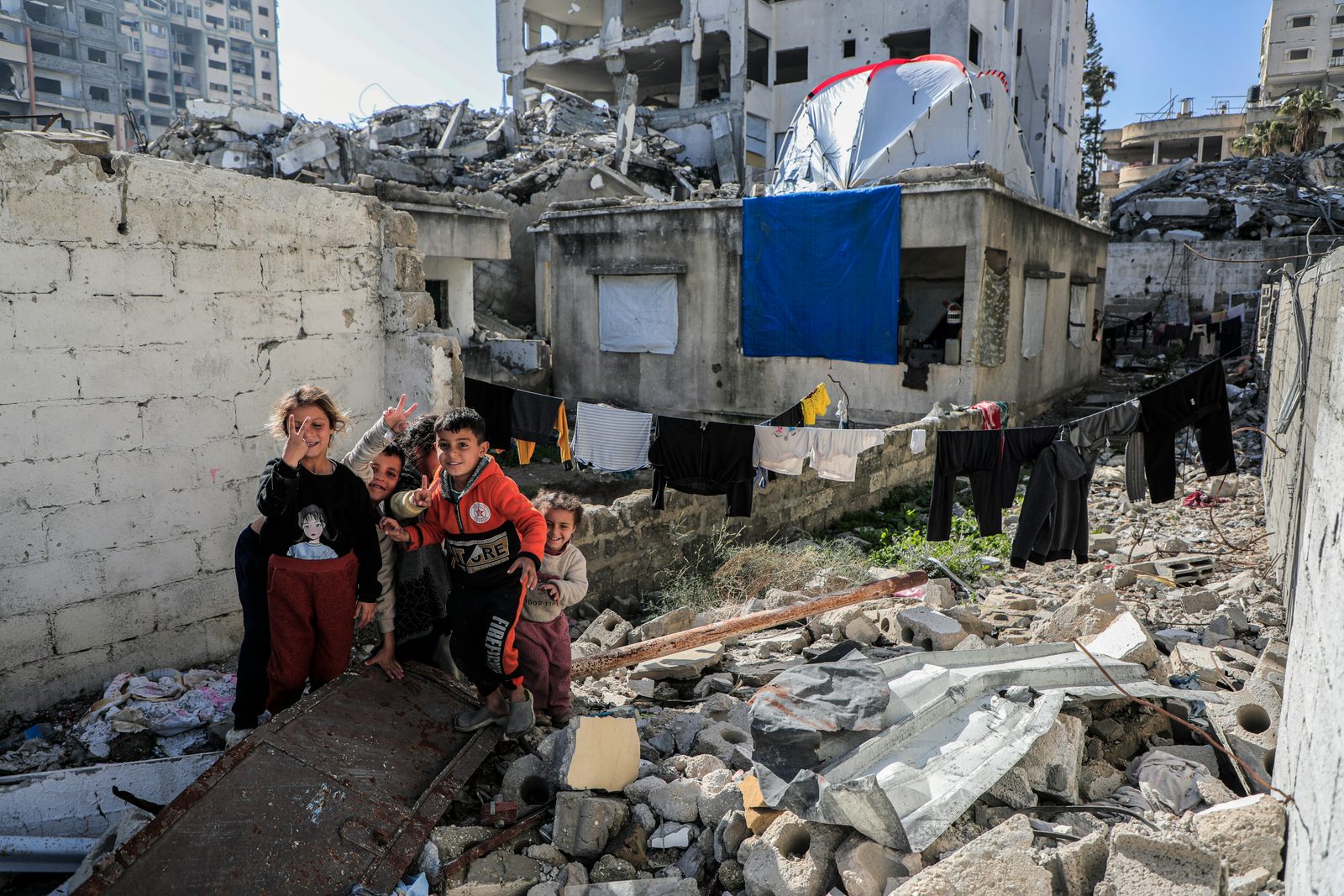 Children stand amid the rubble of destroyed buildings in Gaza, giving peace signs.