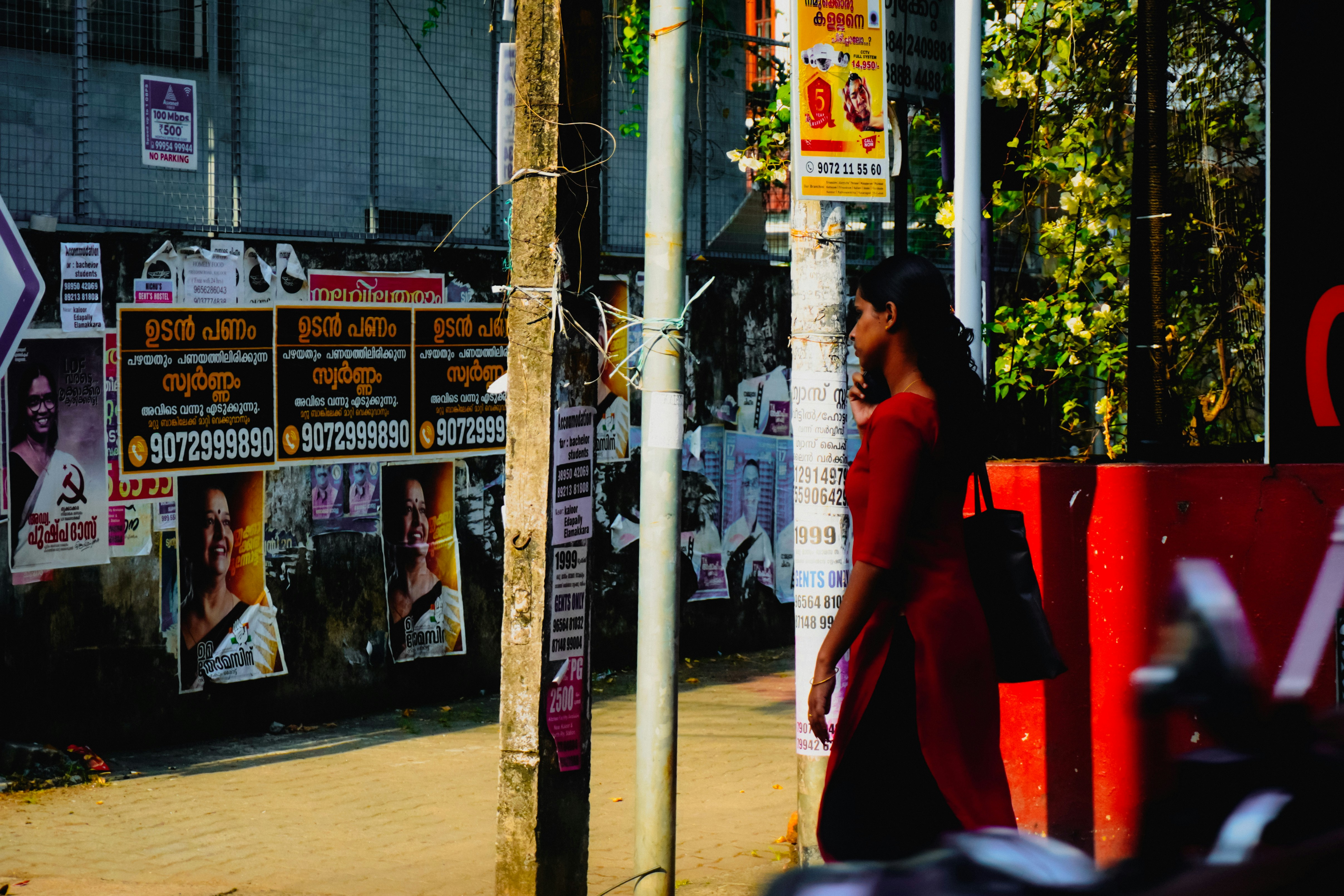 A woman in a red dress walks past a wall covered with political campaign posters in Kerala, India. The posters show candidates from multiple parties, including what appears to be Congress and the Left Democratic Front. Malayalam script is visible throughout. The scene captures everyday street life amid layered political signage.